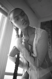 Black and white portrait of a woman with long braided hair wearing a sheer top indoors near a window.