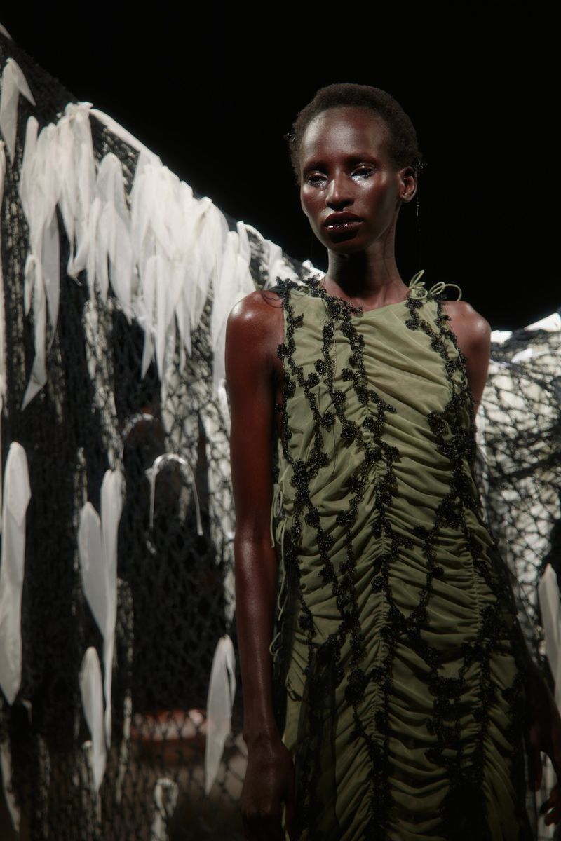 A model on a runway showcases an olive green dress with black lace, accented by a feathered background, in a dimly lit setting.