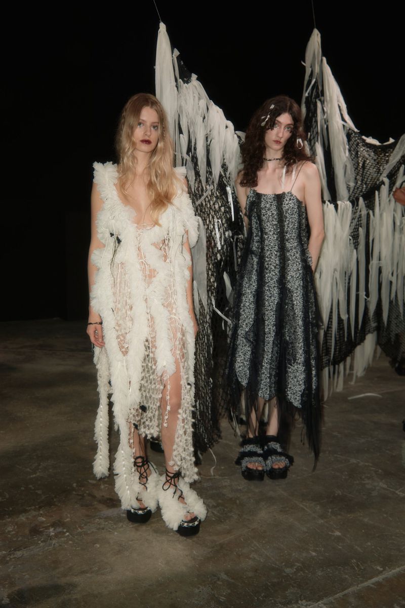 Two fashion models pose in avant-garde dresses with deconstructed fabrics on a concrete floor in a dark studio, showcasing unique designs.
