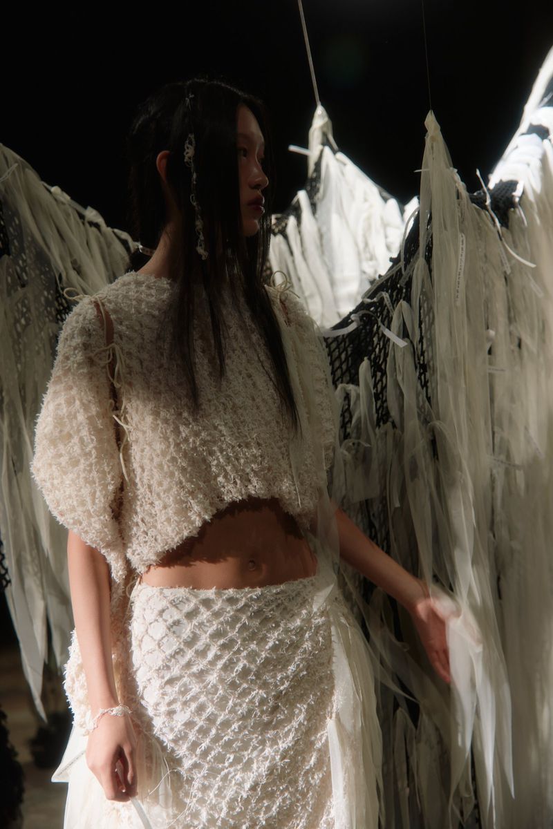 Asian woman poses in a fashion shot wearing a white knitted crop top and skirt with dark hair in an indoor environment with dark lighting.