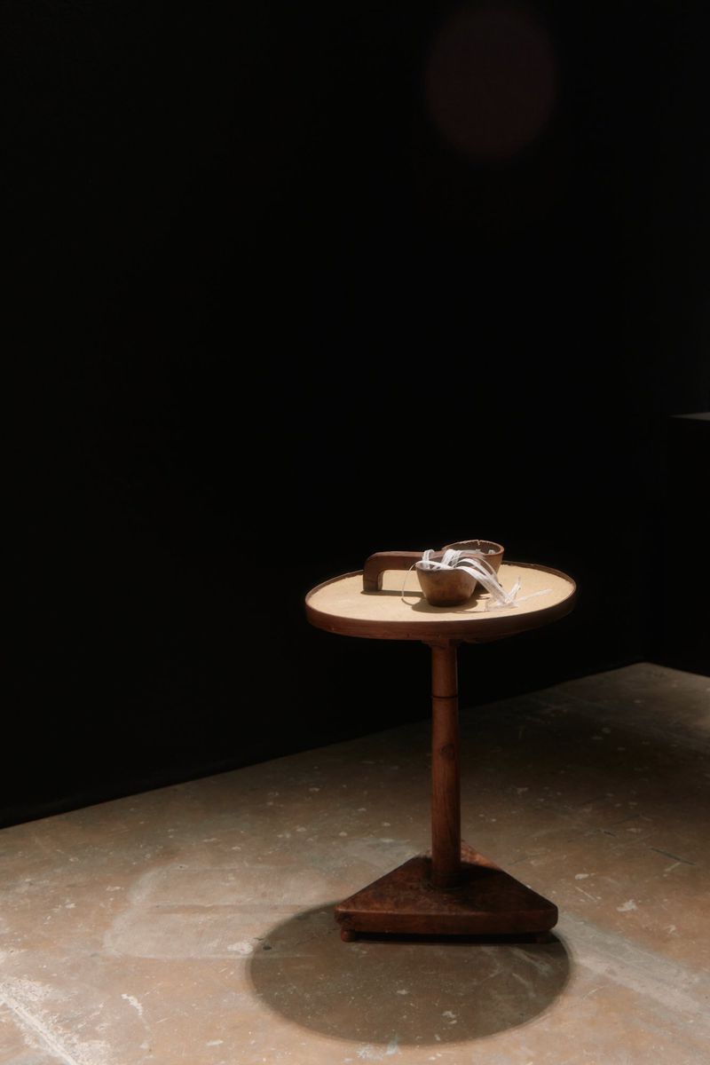 Still life of a tan end table with small wooden bowl and cloth on top, set against a dark background with mottled floor.