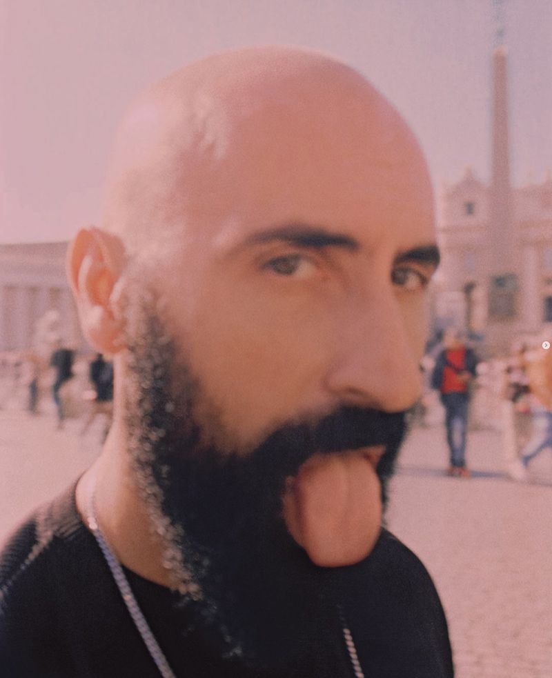 A close-up portrait of a bald man with a dark beard sticking his tongue out in a sunlit plaza in Rome with people and architecture.