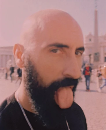 A close-up portrait of a bald man with a dark beard sticking his tongue out in a sunlit plaza in Rome with people and architecture.