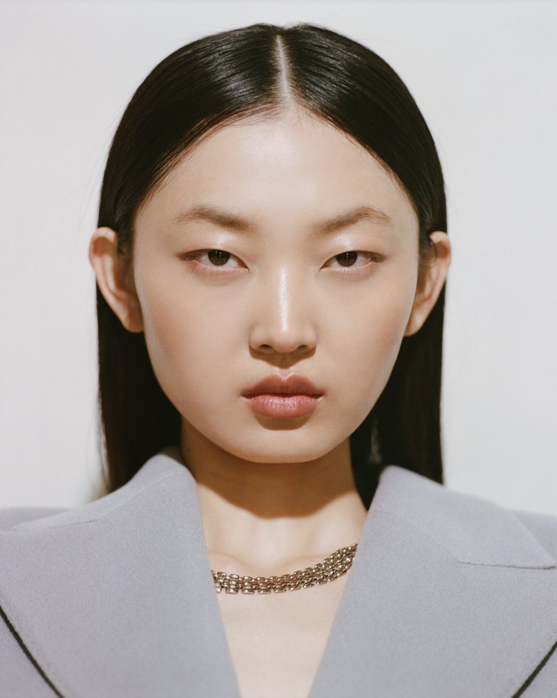 Close-up studio portrait of a serene young adult Asian woman with dark hair, wearing a gray jacket and gold necklace against a white backdrop.