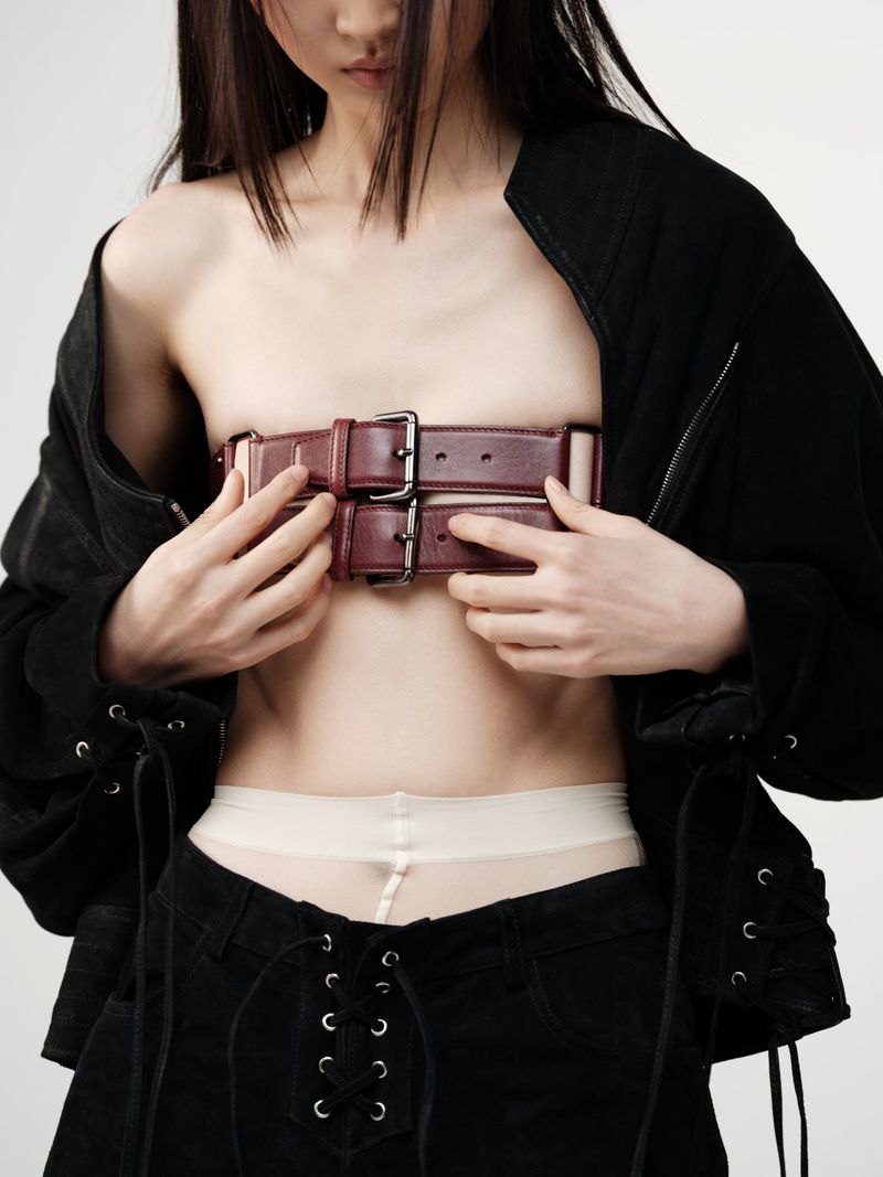 A slender Asian woman poses indoors, wearing black jacket and pants with a pair of leather belts covering her chest against a white backdrop.