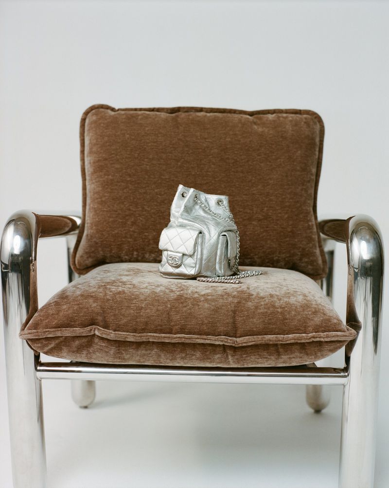 Silver Chanel backpack purse on a brown cushion atop a silver metal frame chair against a plain white backdrop in a studio setting.