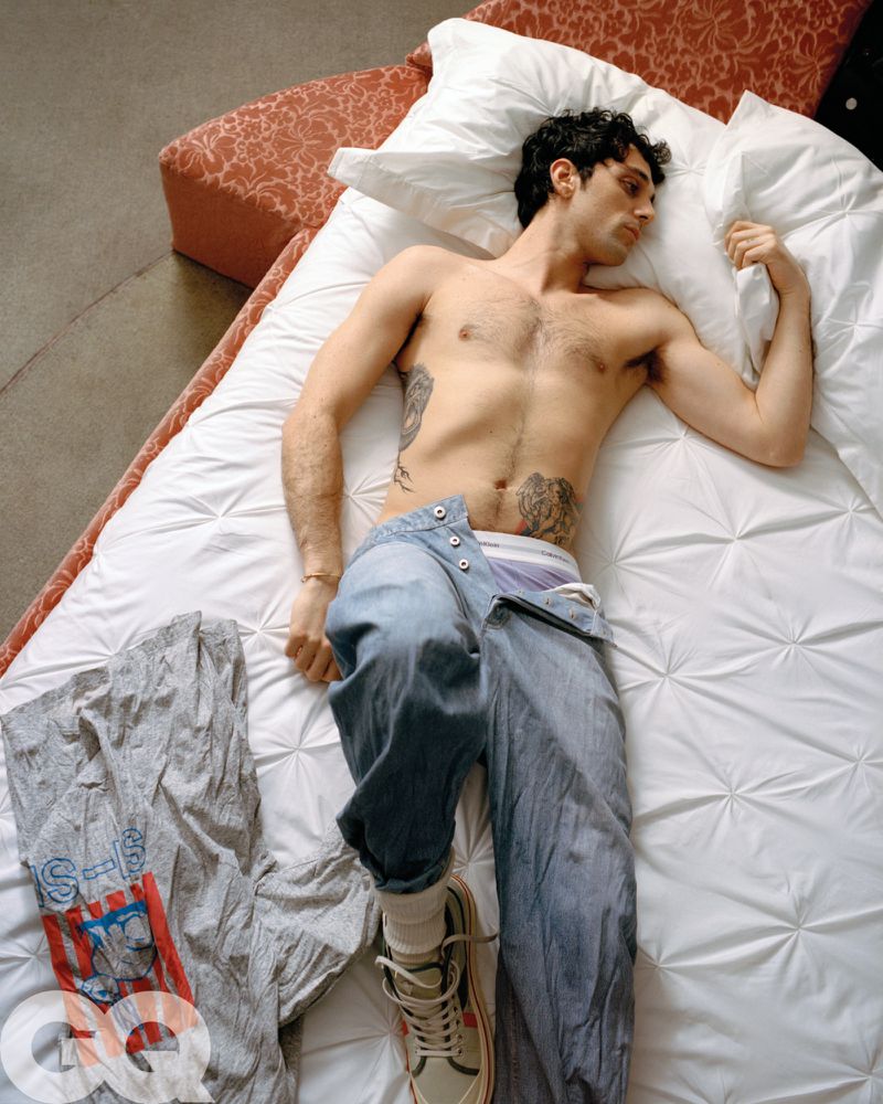 Adult male laying on a white bed with blue jeans, white socks, and sneakers in a light-filled indoor setting with textured concrete floors.