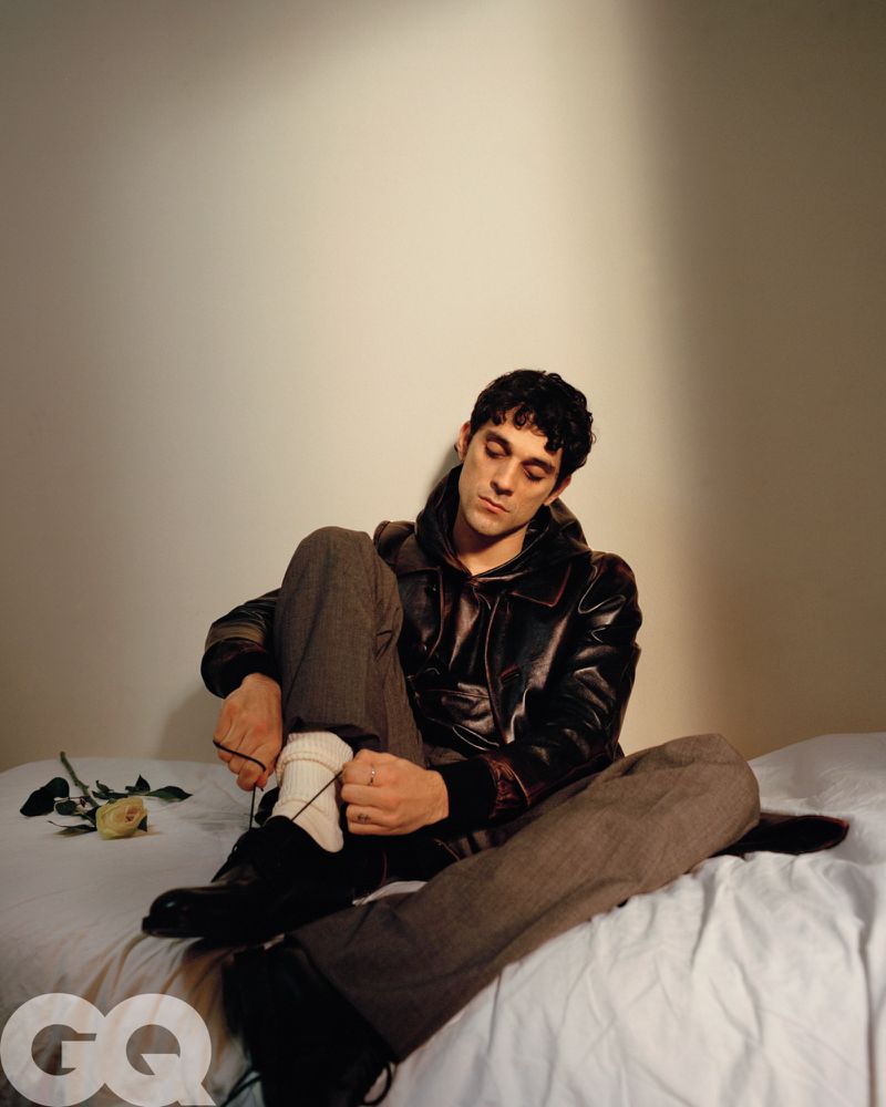 Portrait of a male model sitting on a white bed wearing a leather jacket and grey pants, with a rose on the bed. Indoor lighting.