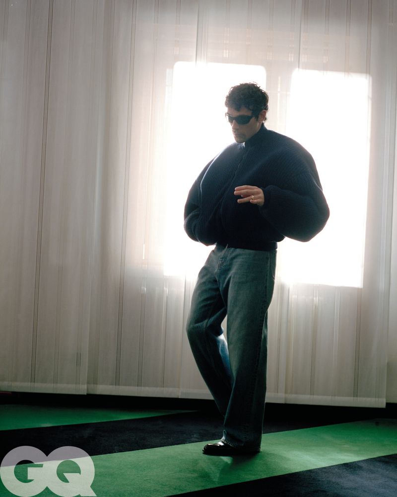A man in a dark jacket and jeans poses in front of a bright window with white curtains and green-and-black flooring, GQ logo in corner.