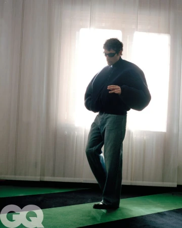 A man in a dark jacket and jeans poses in front of a bright window with white curtains and green-and-black flooring, GQ logo in corner.