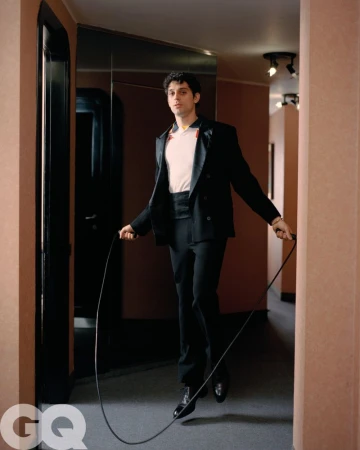 Stylish man in a black suit mid-jump with a jump rope indoors, posing for a GQ magazine photoshoot in a hallway with a neutral color scheme.