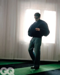 Artist Bresh wearing an oversized dark jacket, sunglasses, and baggy denim jeans standing against backlit sheer curtains.