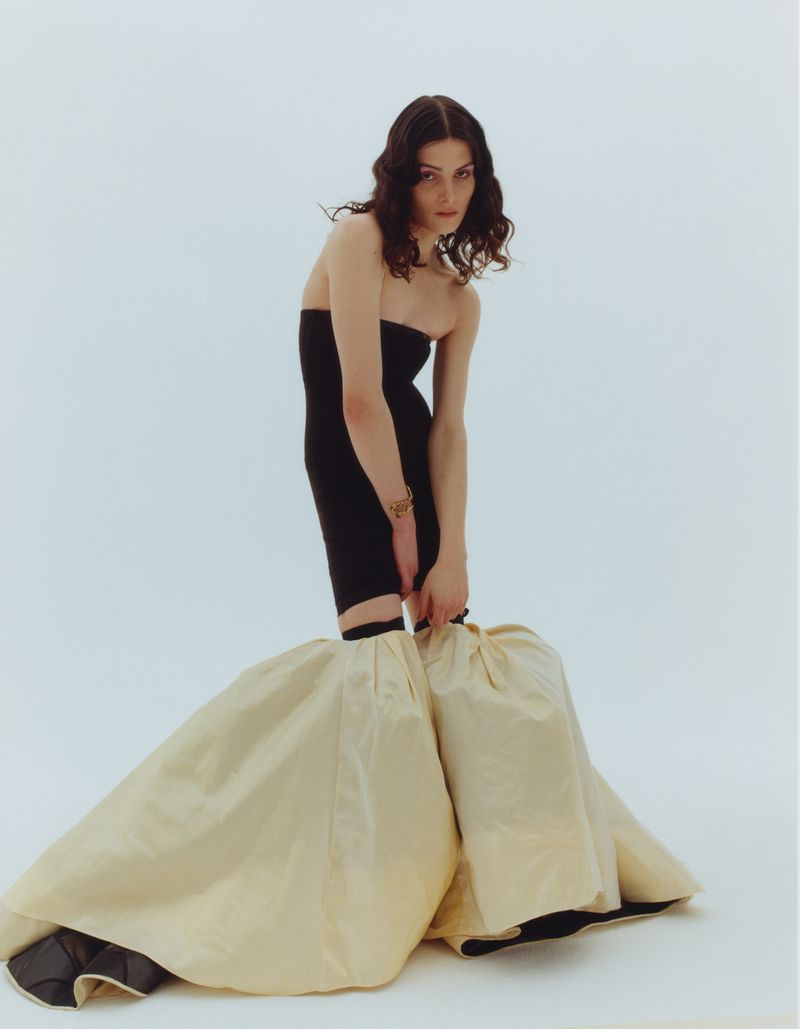 Brunette woman in a black strapless dress with large beige skirt extensions, stands in a white photo studio, looking directly at the camera.