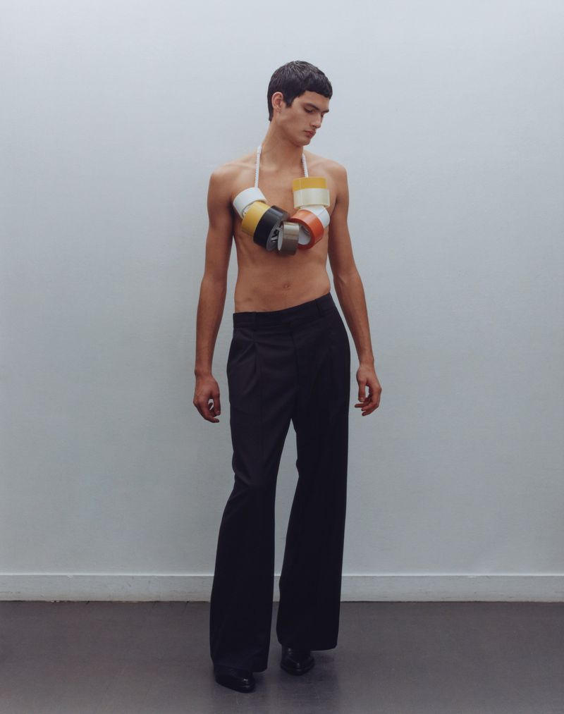 Fashion shot of a young adult male wearing black pants and a unique necklace made of colorful tape rolls in a studio setting.