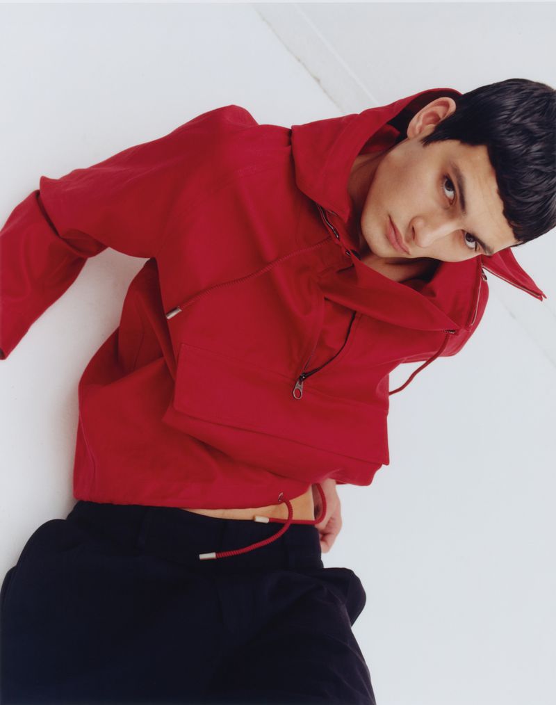 Male model in a vibrant red hooded windbreaker jacket and black pants leaning back against a plain white studio background.