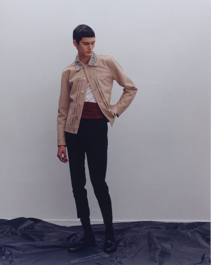A slender man strikes a pose in a tan jacket and black trousers, set against a neutral wall and draped black material.
