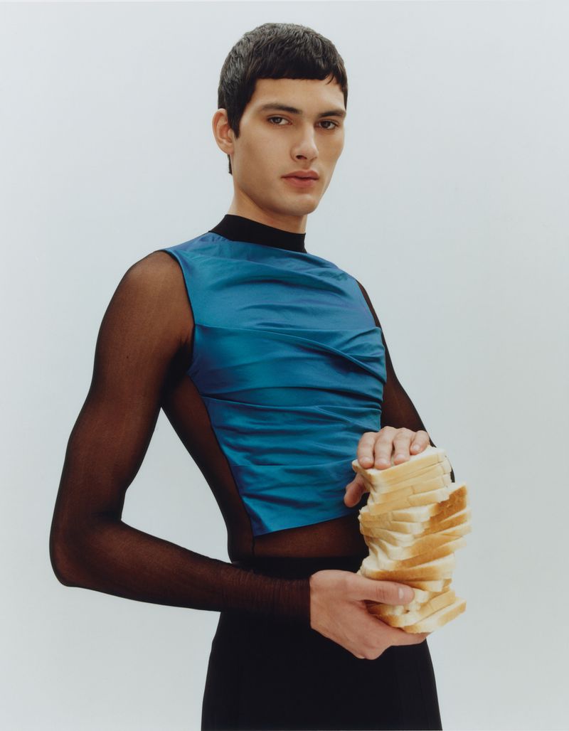 Fashion shot of a young adult male with dark hair, wearing a blue top, holding a vertical stack of white bread slices in a studio setting.