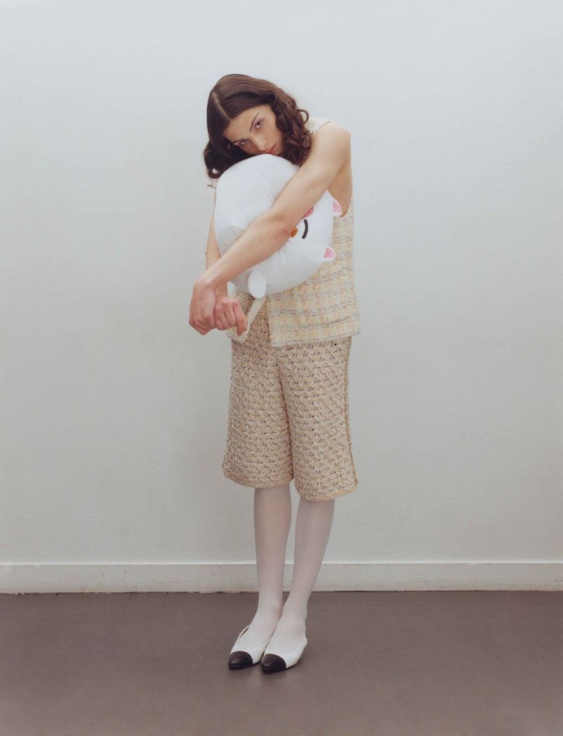 Young woman in a beige patterned outfit hugs a white plush toy against a bright white wall, creating a clean fashion editorial portrait.