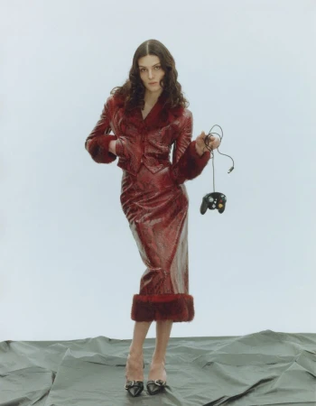 A woman in a red, fur-trimmed suit holds a game controller in a studio. The background is light grey and the floor is textured.