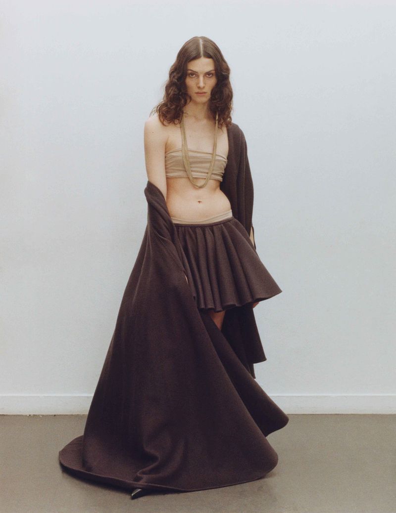 Fashion photo of a young adult woman with wavy brown hair wearing a skirt and tube top, indoors with a neutral background.