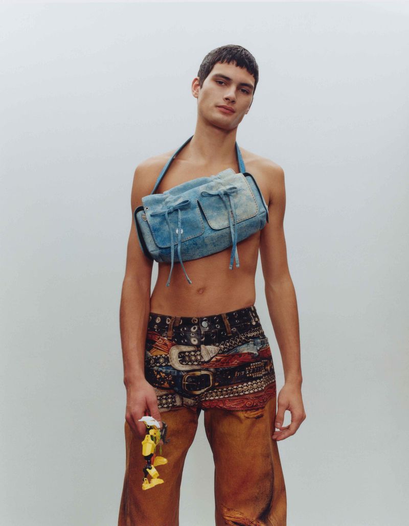 Male model in denim-look shoulder bag and patterned shorts with a yellow object, standing in a neutral studio setting with soft, diffused lighting.