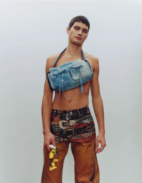 Male model in denim-look shoulder bag and patterned shorts with a yellow object, standing in a neutral studio setting with soft, diffused lighting.