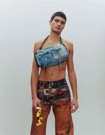 Male model in denim-look shoulder bag and patterned shorts with a yellow object, standing in a neutral studio setting with soft, diffused lighting.