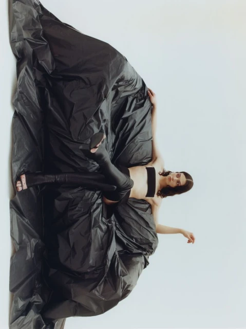 Fashion photo of a woman in a black bandeau and leggings, reclining against a backdrop of black garbage bags in a bright studio.