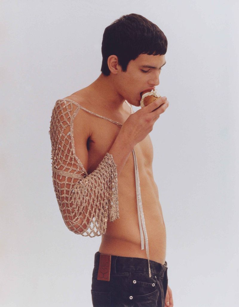 Young adult male eating a pastry indoors. The male is shirtless, wearing jeans and a beaded net-like shawl on one arm.