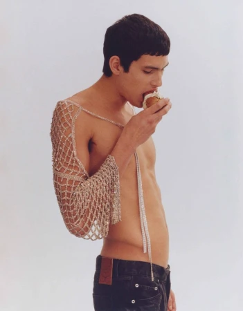 Young adult male eating a pastry indoors. The male is shirtless, wearing jeans and a beaded net-like shawl on one arm.