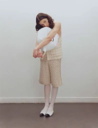 Model wearing a beige tweed vest and matching shorts while clutching a white plush toy against a white studio background.