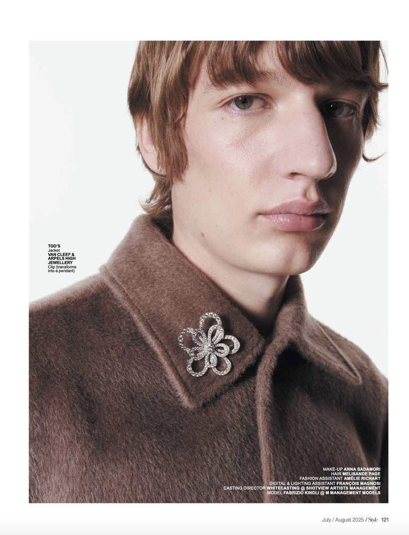 Close-up fashion portrait of a male model with brown hair wearing a brown jacket with a flower brooch against a white backdrop.