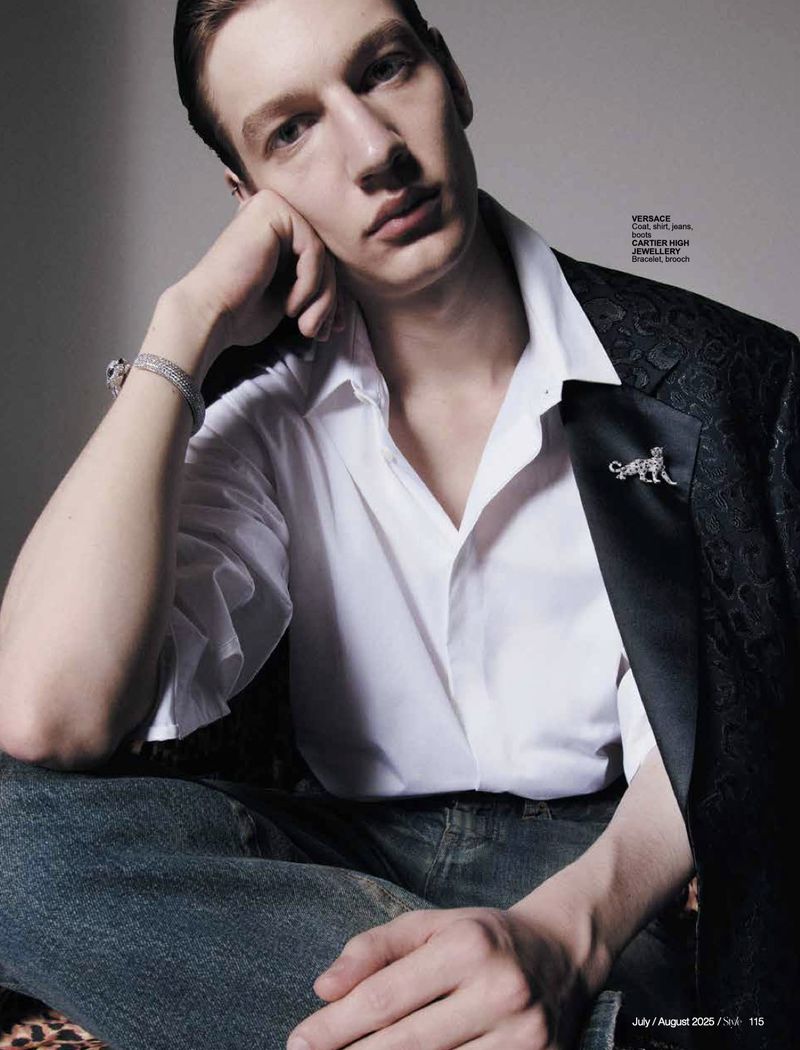 Close-up fashion shot of a young adult male in a white shirt and blazer, seated, accessorized with bracelets and a pin, in a studio setting.