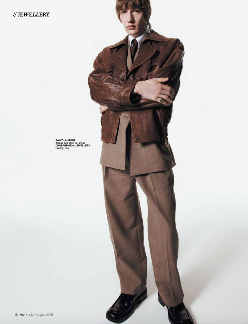 Man poses in Saint Laurent brown leather jacket, brown suit, and black dress shoes in a brightly lit photo studio.