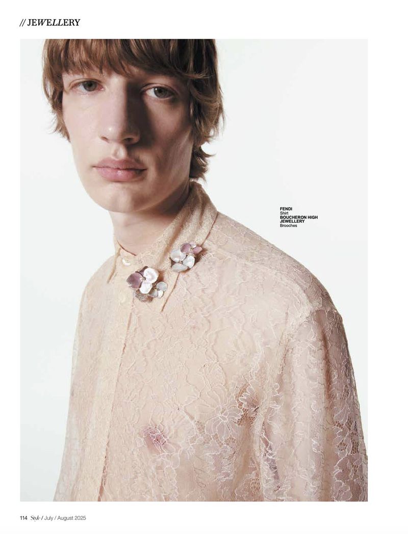 Headshot of a young adult man wearing a peach-colored lace button-down shirt, accessorized with two floral brooches on the collar.