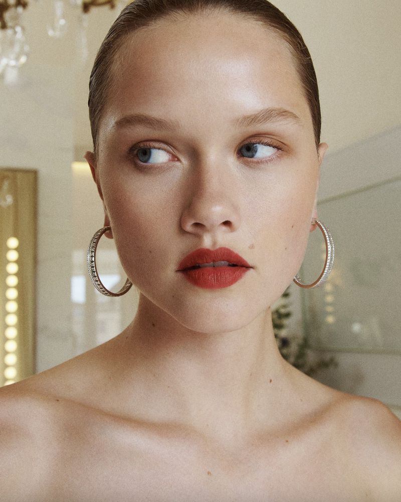 Close-up portrait of a young adult woman with fair skin and red lipstick, wearing hoop earrings and looking to the side in an indoor setting.