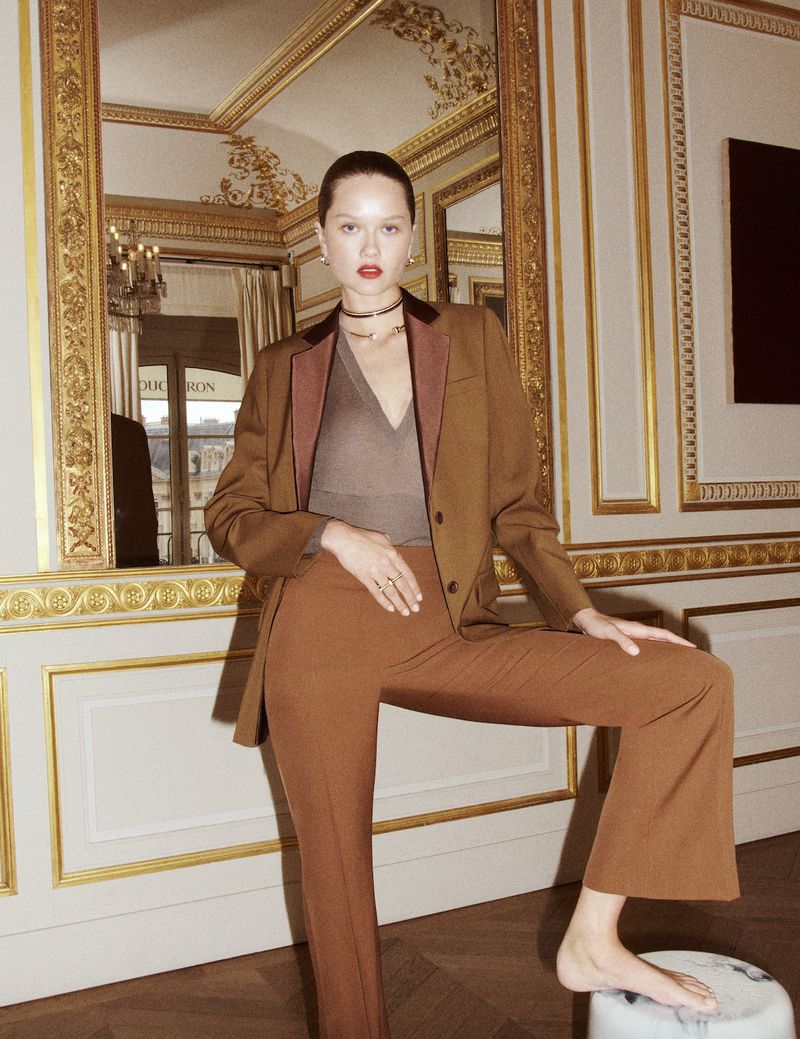 Stylish woman in a brown blazer and matching trousers poses indoors with one leg on a marble stool, against a decorative wall.