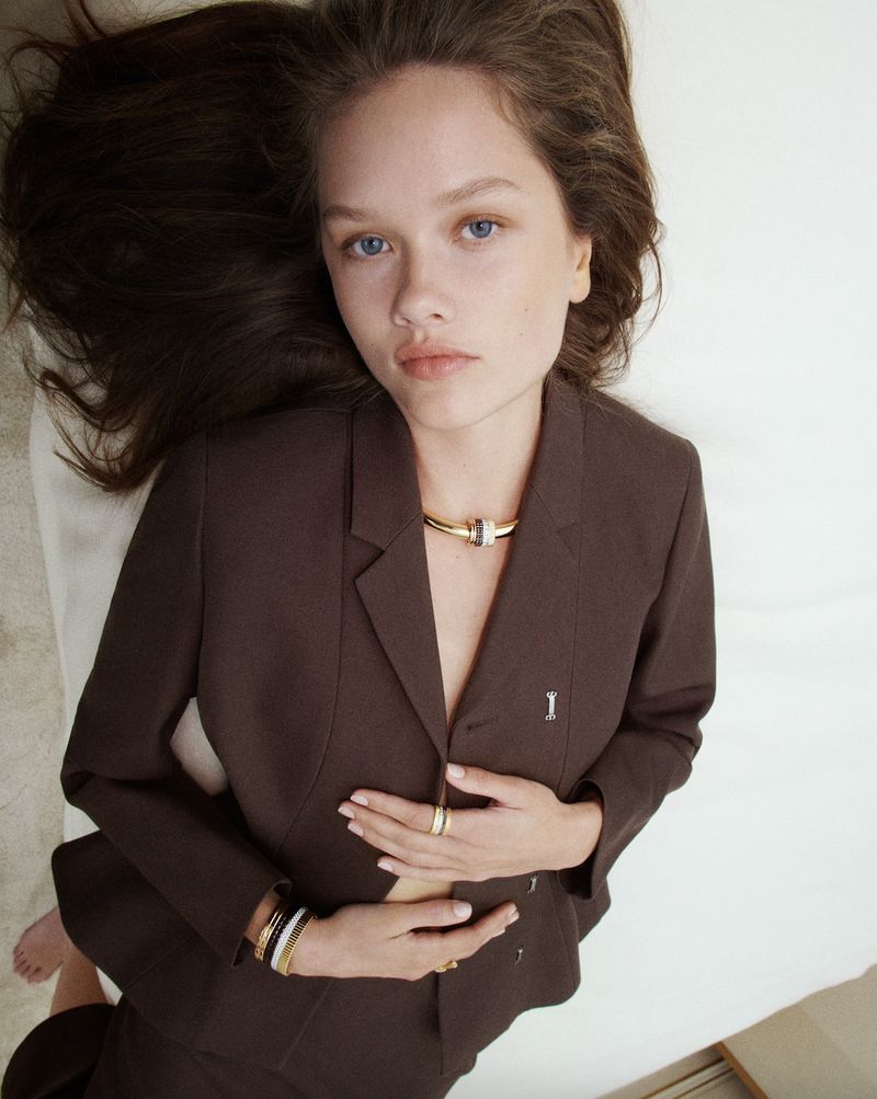 Woman in a brown suit lying on a white surface, wearing a gold collar necklace and bracelets, with long brown hair framing her face.