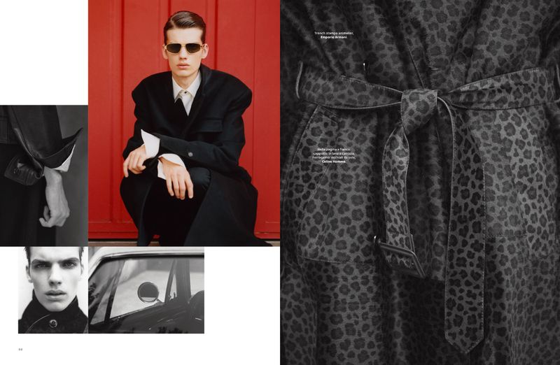 Fashion editorial image: a male model wearing sunglasses and a black overcoat poses in front of a red door with a grey car in the background.