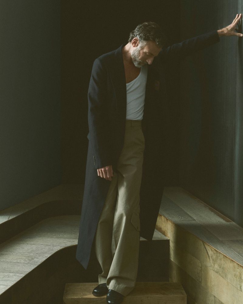 Mature man in a dark coat and light pants leans against a dark wall, standing on wooden steps in soft, diffused lighting indoors.