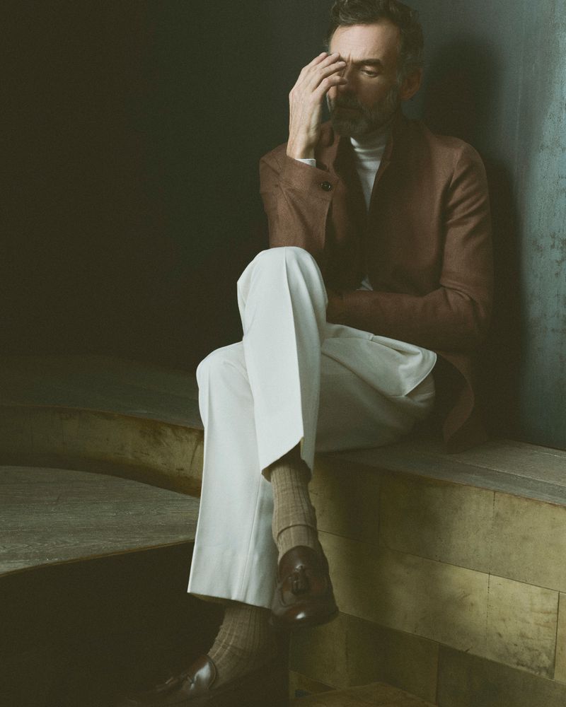 Portrait of mature adult man in brown suit and white pants sitting indoors with pensive expression, fashionable loafers and neutral backdrop.