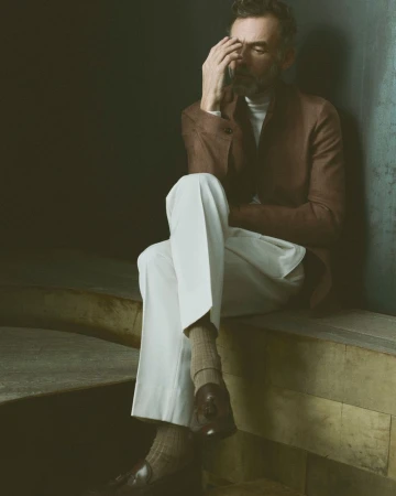 Portrait of mature adult man in brown suit and white pants sitting indoors with pensive expression, fashionable loafers and neutral backdrop.