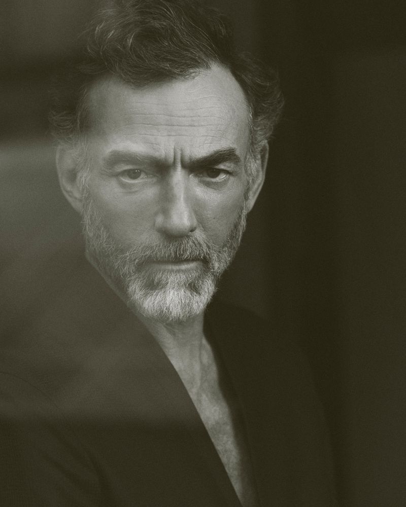 Portrait of an adult male with grey hair and beard looking directly at the camera wearing a dark jacket against a dark backdrop.