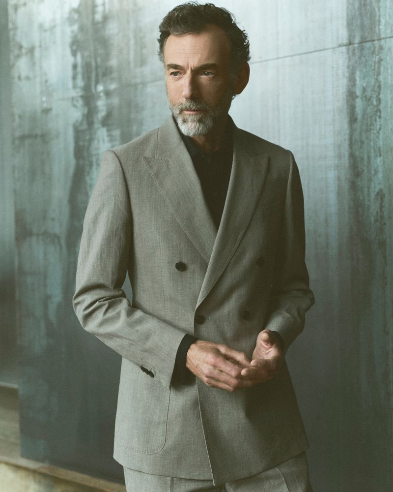 Portrait of an adult male with a beard wearing a light grey double-breasted suit in front of a textured grey wall background.