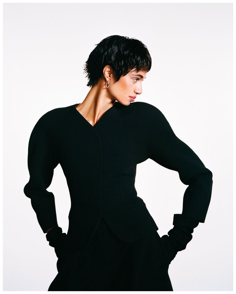 Fashionable woman with short dark hair poses in a black outfit with exaggerated shoulders against a bright white backdrop in a studio setting.