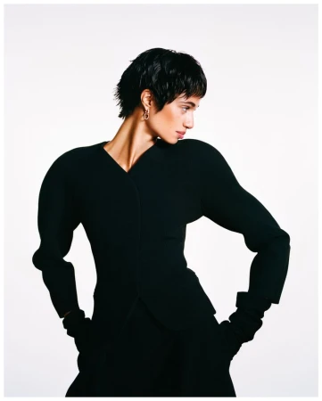 Fashionable woman with short dark hair poses in a black outfit with exaggerated shoulders against a bright white backdrop in a studio setting.