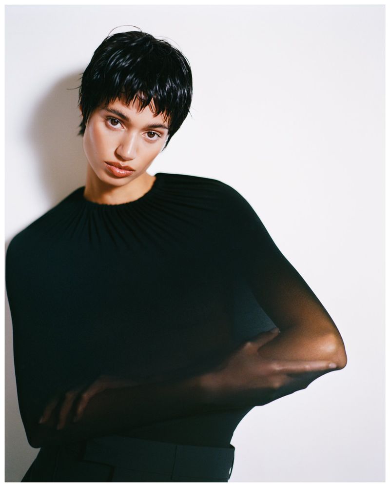 Model Malika El Maslouhi posing with arms crossed in a sheer black top against a bright white background.