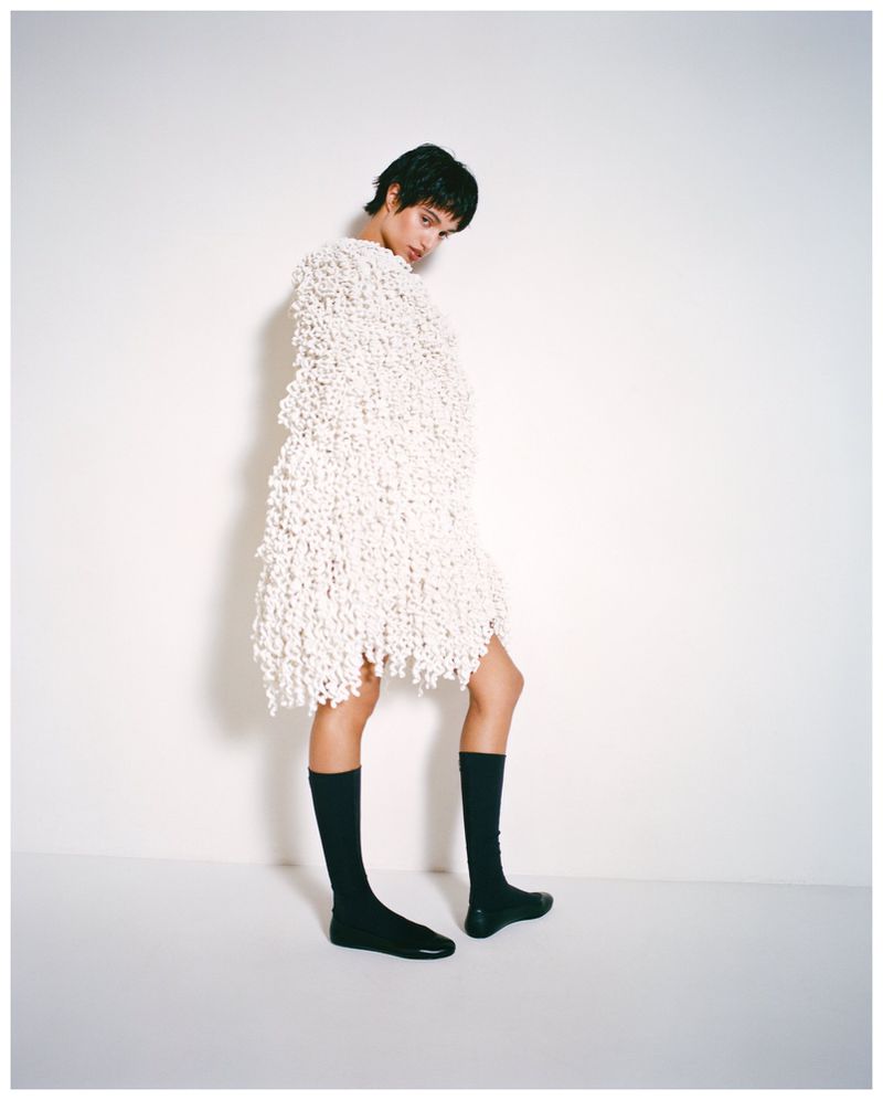 Model Malika El Maslouhi wearing a textured white knit cape dress and black knee-high socks against a white studio background.