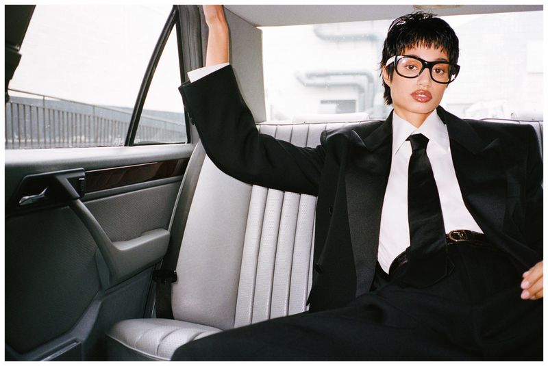 A young adult woman with short hair, wearing glasses and a black suit with a tie, posing inside a car with grey seats, against a blurred urban backdrop.