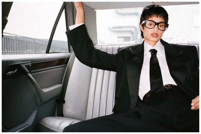 A young adult woman with short hair, wearing glasses and a black suit with a tie, posing inside a car with grey seats, against a blurred urban backdrop.